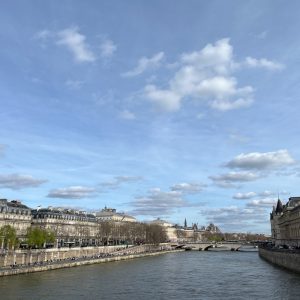 Vue d'un ciel bleu parsemé de nuages au-dessus de la Seine à Paris, avec des bâtiments historiques et un pont en arrière-plan.